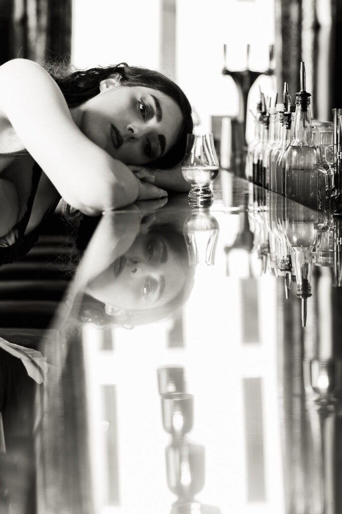 Absente Présence est un portrait en noir et blanc d’une femme perdue dans ses pensées, front posé sur un bar, face à un verre vide. Son reflet flou évoque l’attente, le manque, peut-être un rendez-vous manqué.