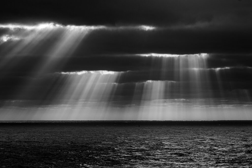 Rébellion Lumineuse est une photographie en noir et blanc saisissante issue de la série &quot;Keshiki&quot;, capturant un moment unique où le soleil perce les nuages sombres au-dessus de la Manche, révélant une scène dramatique de lumière contre ténèbres.