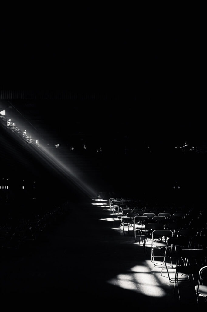 Photographie d&rsquo;art en noir et blanc montrant une salle vide avec des chaises align&eacute;es, travers&eacute;e par une lumi&egrave;re rasante, intitul&eacute;e Entre deux Silences par Art Par Sten.