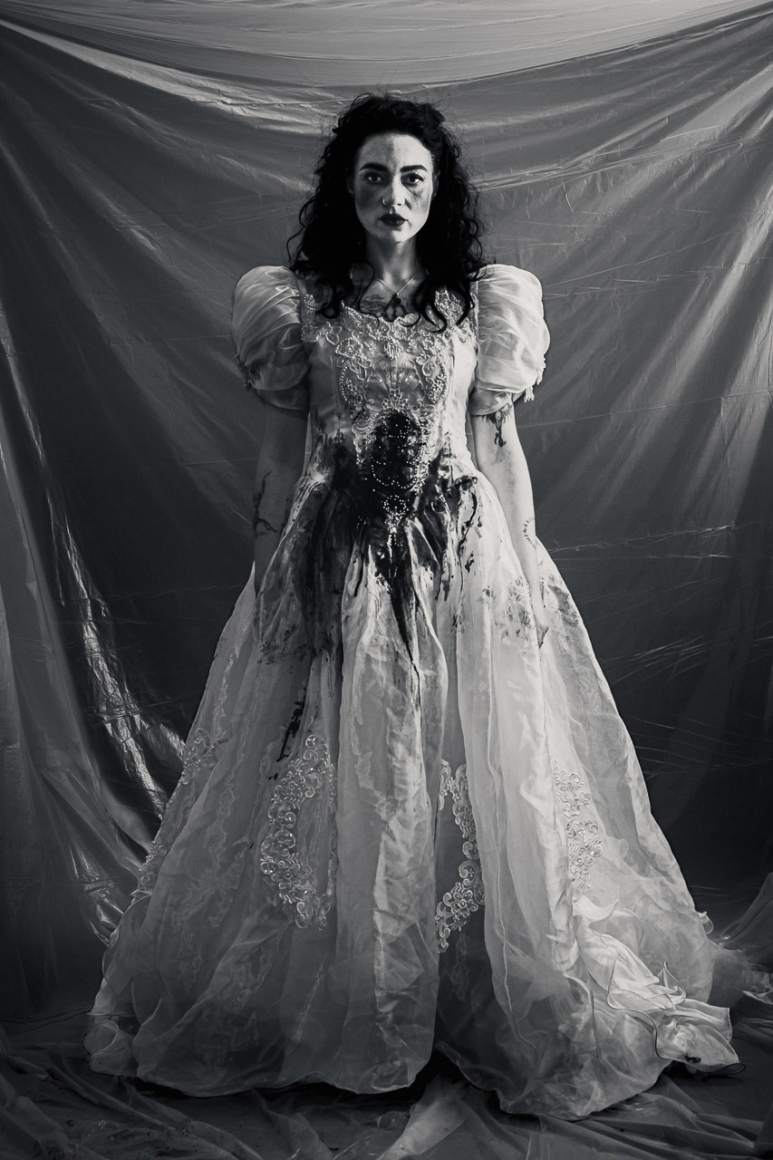 Portrait artistique en noir et blanc d’une femme debout en robe blanche, regard face caméra, robe tachée de noir, fond drapé.