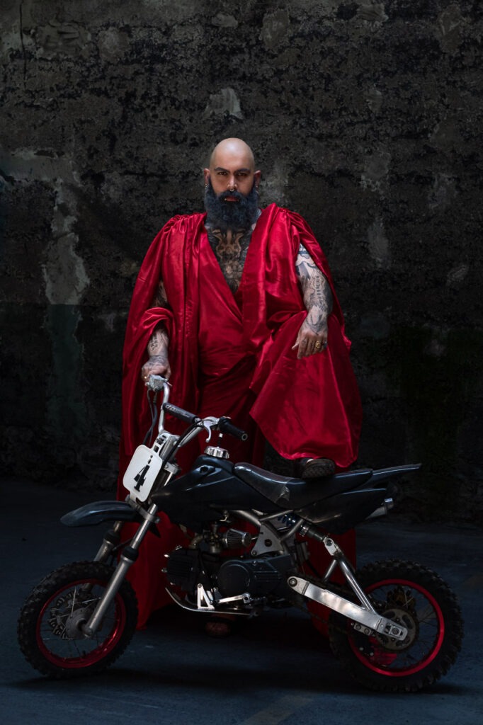 Empire Minuscule de Thierry Stentzel, photographie d&rsquo;art d&rsquo;un homme tatou&eacute; en cape rouge, une jambe pos&eacute;e sur une mini-moto dans un d&eacute;cor sombre.