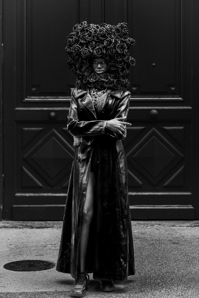 Reine Debout Reine Debout de Thierry Stentzel, photographie d’art noir et blanc, portrait d’une femme noire coiffée d’une imposante parure de roses devant une porte monumentale.